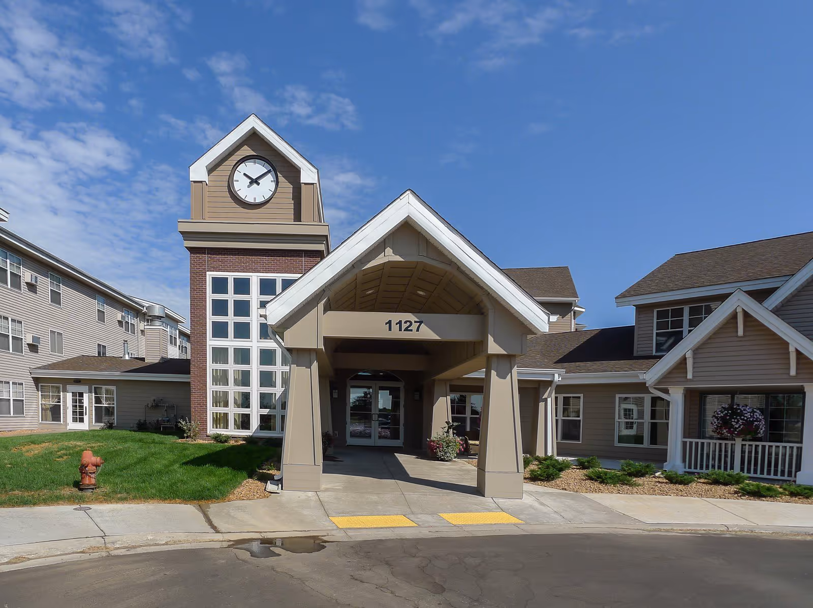 Front entrance of a senior living building with a covered porte-cochere, clock tower, and address number 1127.