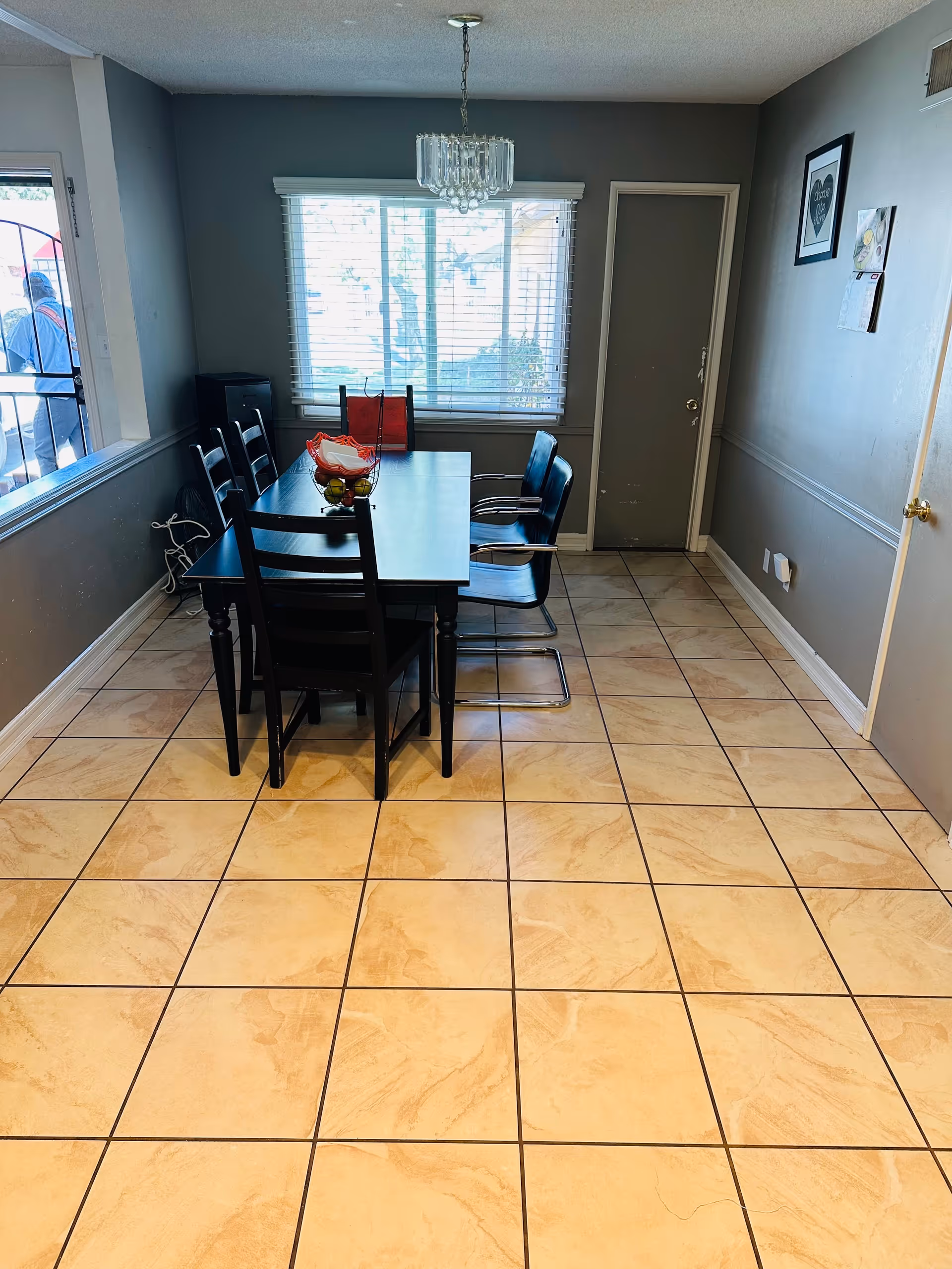 Rectangular dining table with chairs on a tiled floor under a chandelier in a small dining room with a window.