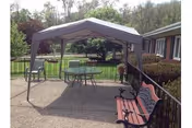 Outdoor patio area with a canopy-covered table and chairs, a bench, and railing overlooking a grassy yard and trees.