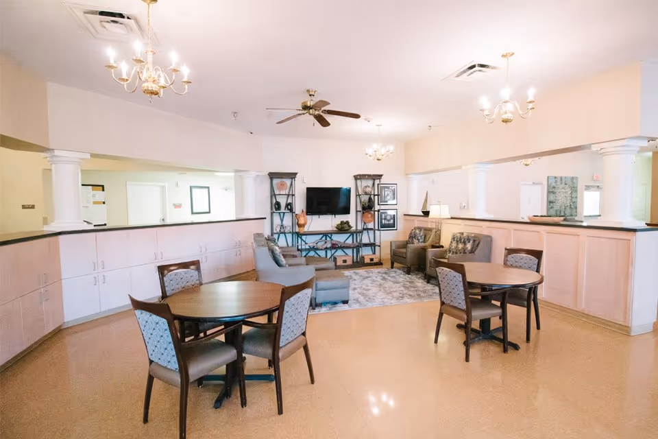 A spacious senior living facility common area with round tables, upholstered chairs, couches, and a television.