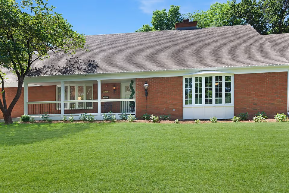 Exterior view of a single-story brick building with a gray shingled roof, a white porch railing, a bay window, and a well-maintained green lawn in front. There is a tree on the left side and some small shrubs along the building's foundation.