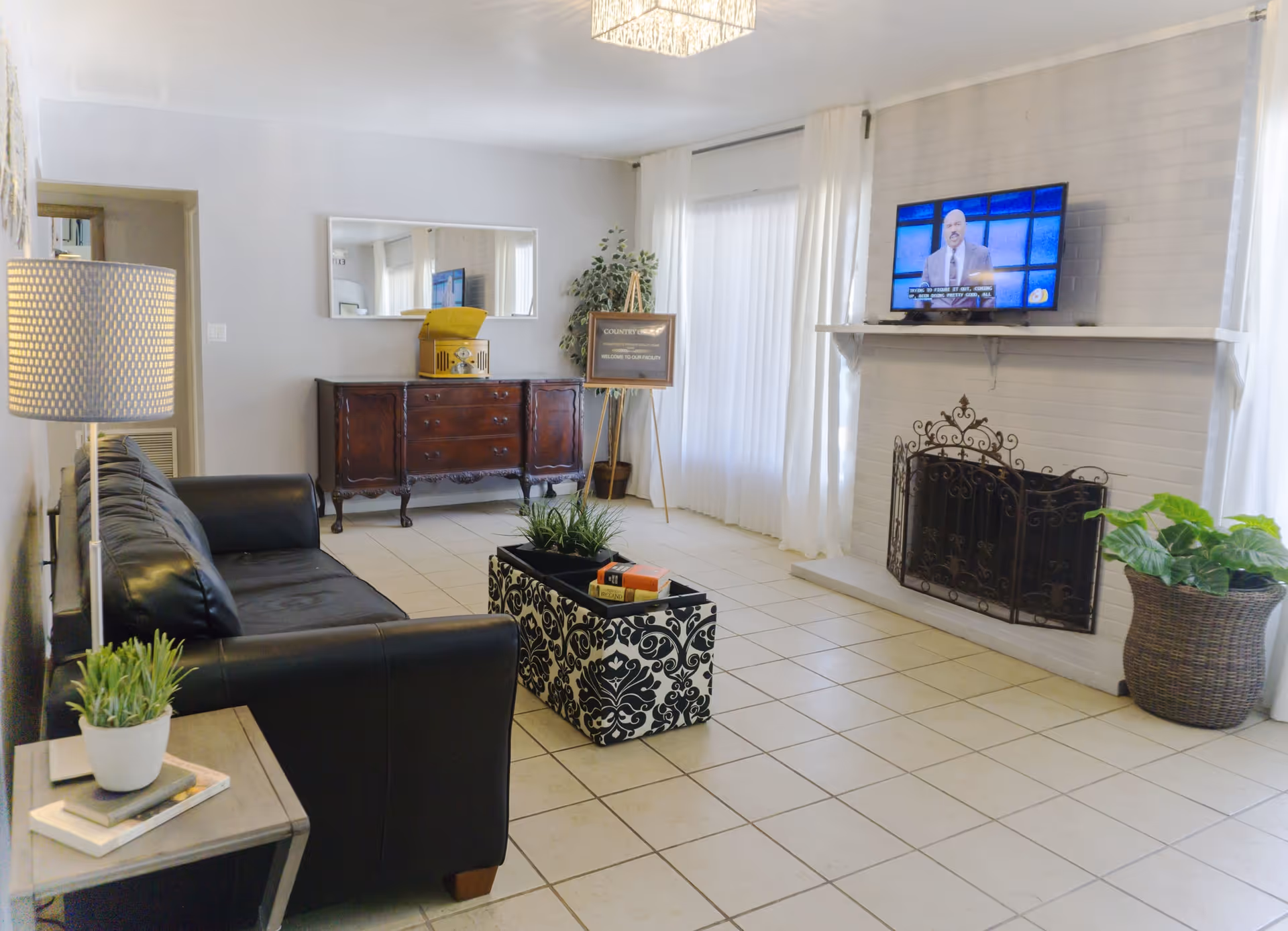 A bright living room with a black leather sofa, a patterned coffee table with books and plants, a wooden sideboard with a mirror above it, a fireplace with a decorative screen, a wall-mounted TV, and large windows with white curtains letting in natural light.