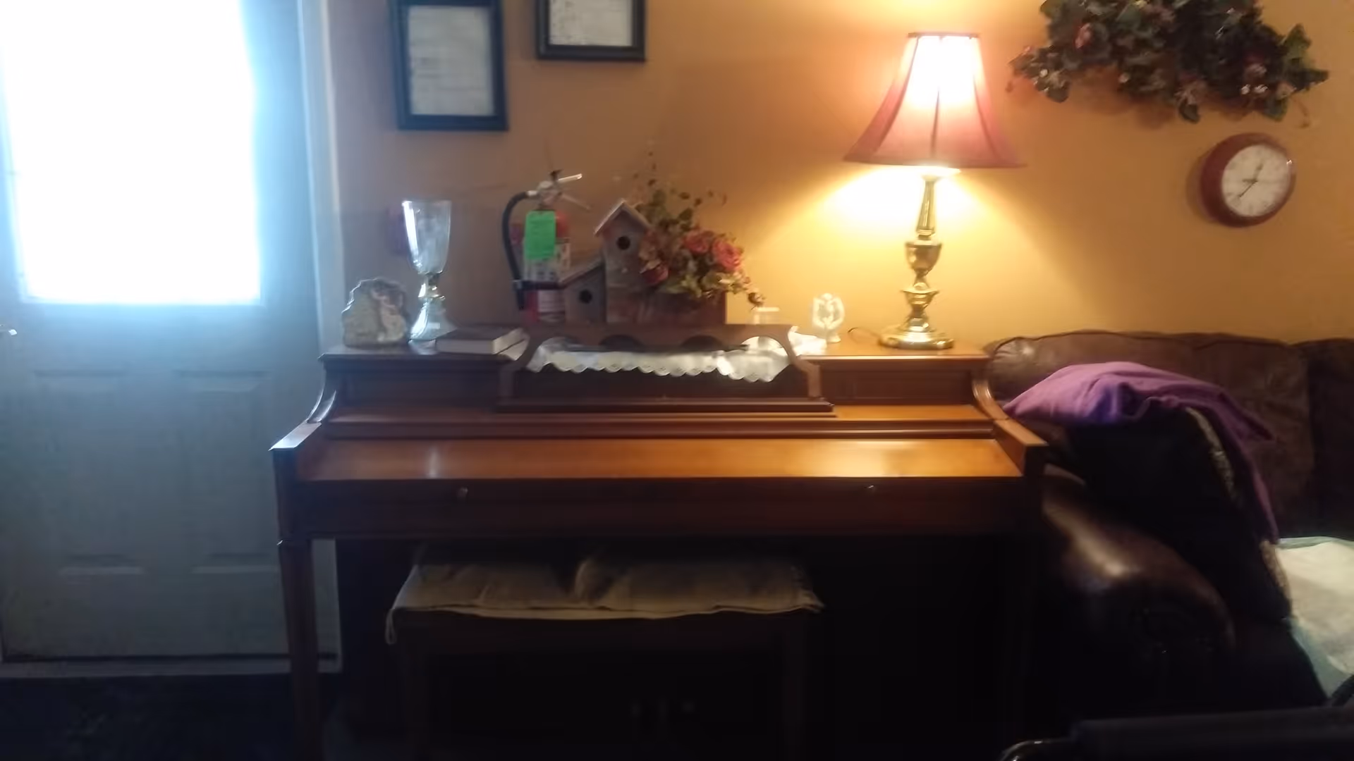 Upright wooden piano against a wall with decorative items and a table lamp, next to a leather couch and a front door.