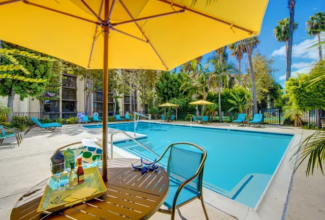 Outdoor swimming pool area at Meridian at Laguna Hills with several lounge chairs and tables with yellow umbrellas. A round table in the foreground holds a tray with bottles and glasses. The pool is surrounded by greenery and palm trees under a clear blue sky.