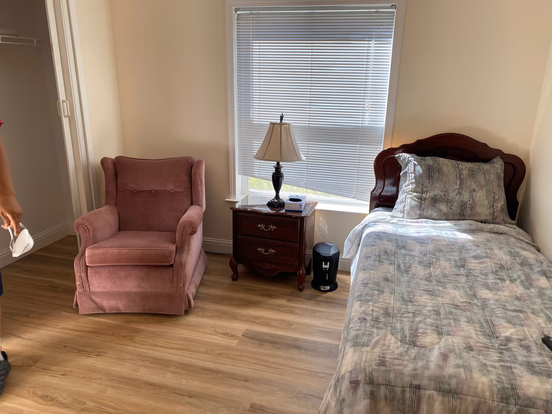 A small bedroom with a single bed covered in a patterned comforter and pillow, a wooden nightstand with a lamp and a small box on it, a pink upholstered armchair, and a window with closed blinds letting in some light. Part of a person holding a face mask is visible on the left side.