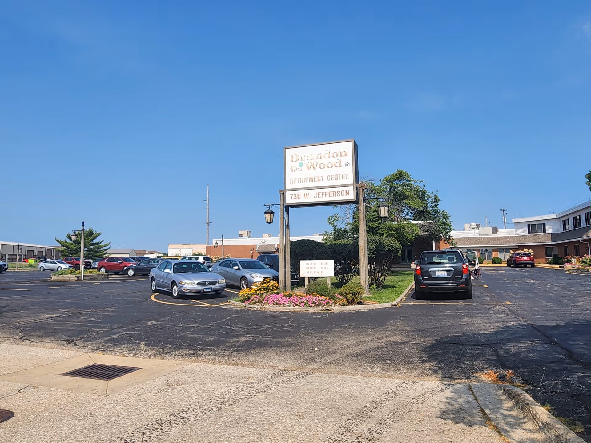 Parking lot area in front of Brandon Wood Retirement Center with several parked cars, a sign displaying the facility name and address, and a clear blue sky.