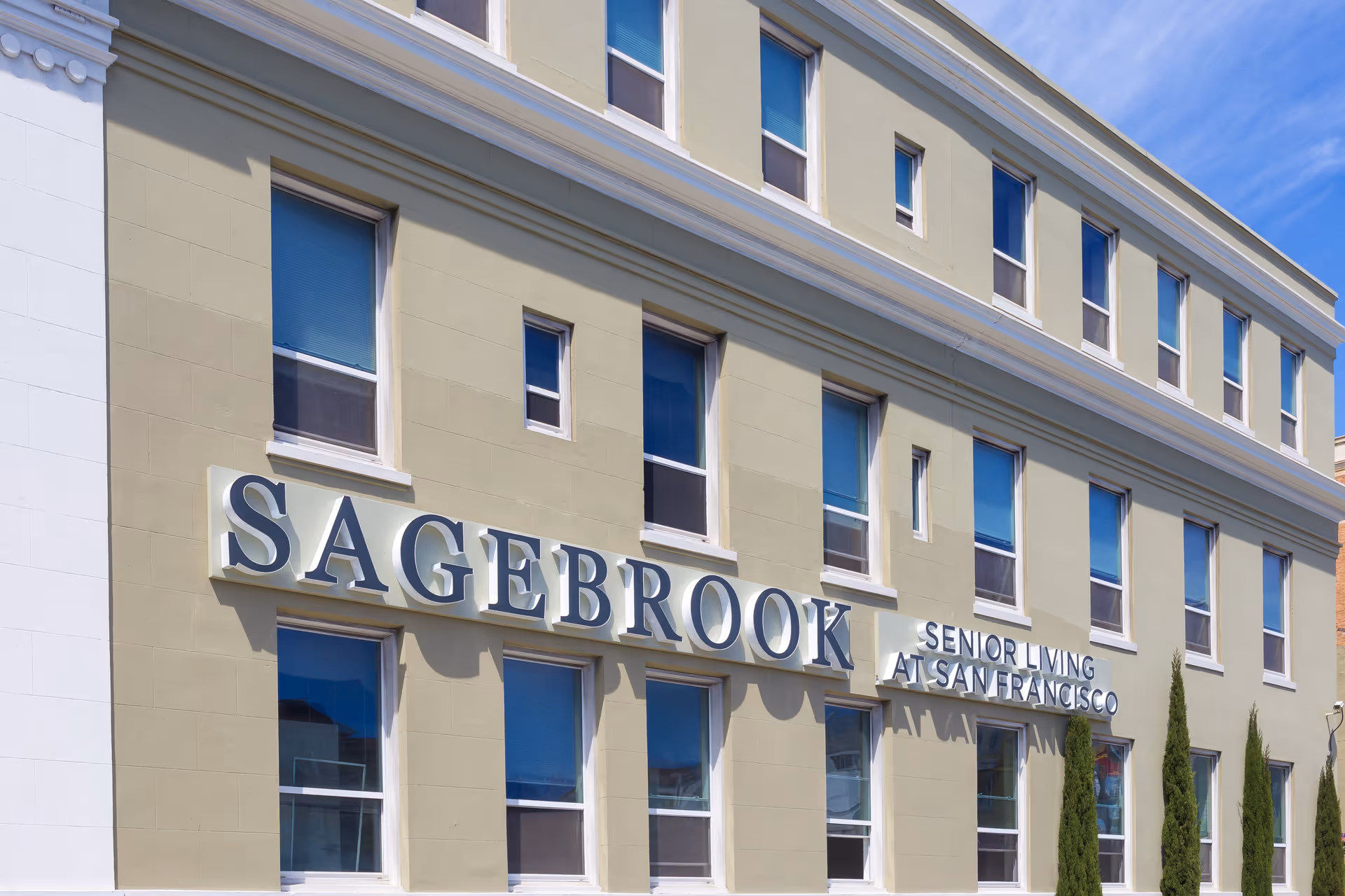 Exterior view of a multi-story building with beige walls and multiple windows. The building has a large sign that reads 'SAGEBROOK SENIOR LIVING AT SAN FRANCISCO'. There are a few tall, narrow trees planted in front of the building under a clear blue sky.