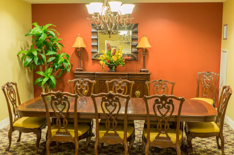A formal dining room with a long wooden table surrounded by eight ornate wooden chairs with yellow cushions. Behind the table is a wooden sideboard with two lamps and a floral arrangement in the center. A decorative mirror hangs on the red accent wall above the sideboard. A large green potted plant is placed in the corner to the left.