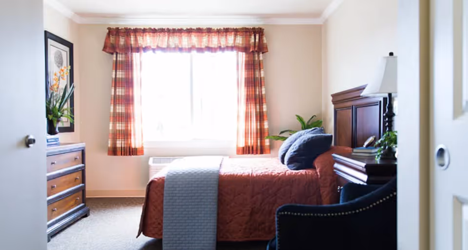 A cozy bedroom with a bed covered in a rust-colored quilt and a light blue throw blanket. The bed has two blue pillows and a wooden headboard. Next to the bed is a wooden nightstand with a lamp and some books. A dark blue armchair is partially visible in the foreground. A window with red and orange plaid curtains lets in natural light. A wooden dresser with a plant and framed artwork is on the left side of the room.