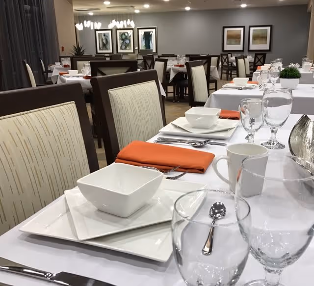 A dining room set up with tables covered in white tablecloths, each place setting featuring white square plates, bowls, silverware, clear glassware, white coffee cups, and orange cloth napkins. The room has beige upholstered chairs with dark wooden frames, framed artwork on the walls, and soft overhead lighting.