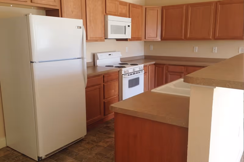 A kitchen with wooden cabinets, a white refrigerator, a white microwave mounted above a white electric stove, and a countertop with a sink. The floor has a patterned tile design.
