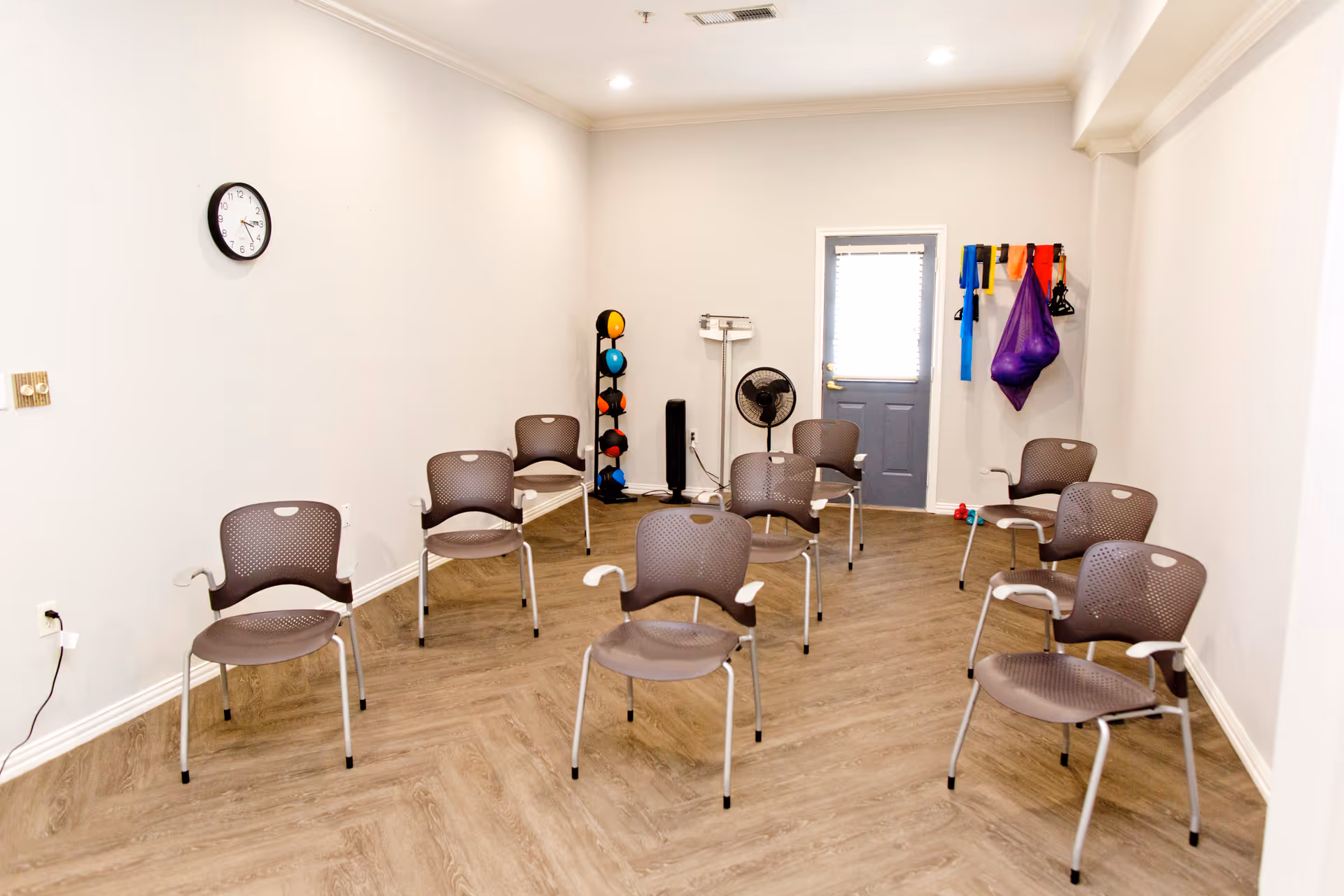 A small exercise or therapy room with eight brown chairs arranged in a spaced-out pattern on a wood-patterned floor. On the back wall, there is a rack holding colorful exercise balls and resistance bands, a standing fan, and a scale. A clock is mounted on the left wall, and a door with a window is at the back of the room.