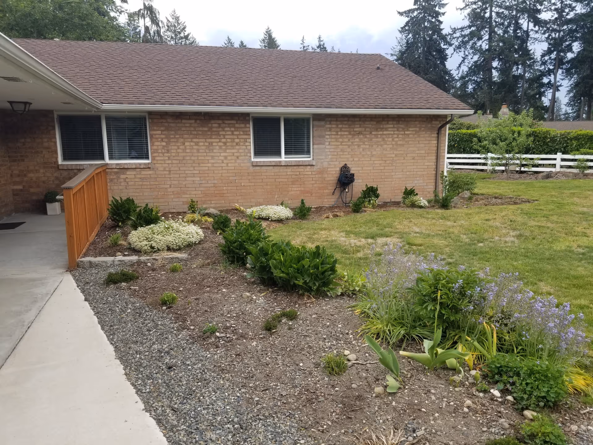 Outdoor view of a single-story brick building with two windows, a brown shingled roof, and a garden bed with various green plants and flowers in front. There is a concrete walkway on the left side and a grassy lawn on the right, with trees and a white fence in the background.