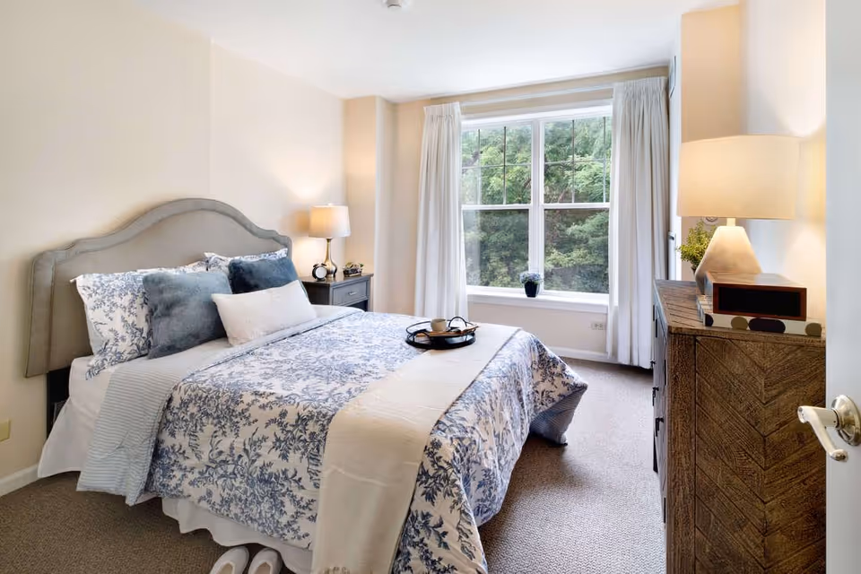 A bright and cozy bedroom with a large window showing green trees outside. The bed has a gray upholstered headboard and is dressed with white and blue floral bedding, multiple pillows, and a beige throw blanket. There is a nightstand with a lamp and clock on one side of the bed, and a wooden dresser with a lamp and decorative items on the other side. White curtains frame the window, and slippers are placed neatly on the carpeted floor near the bed.