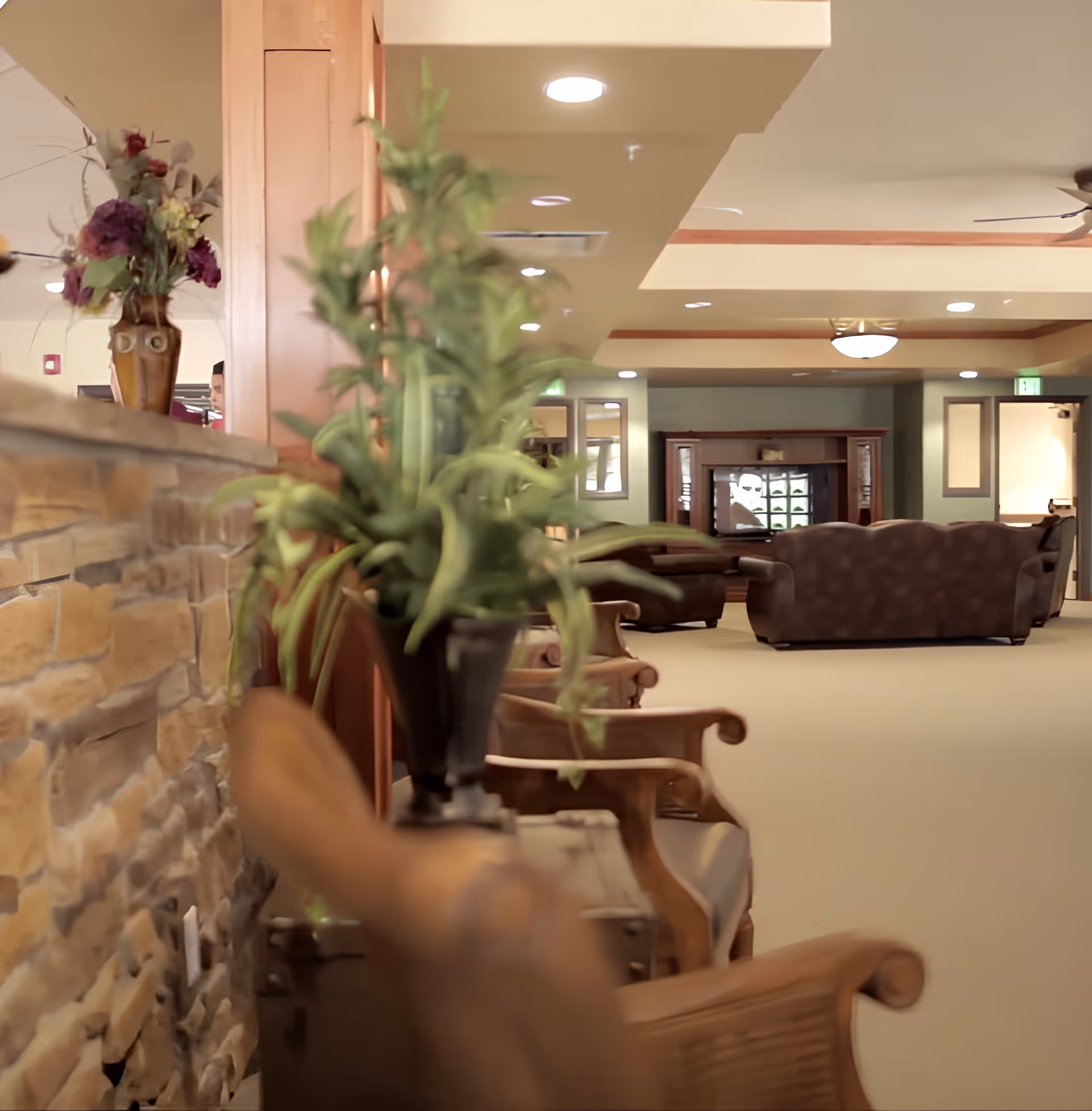 Interior view of a senior living facility lounge area with wooden chairs, a stone wall, potted plants, and a seating area with sofas and a television in the background.