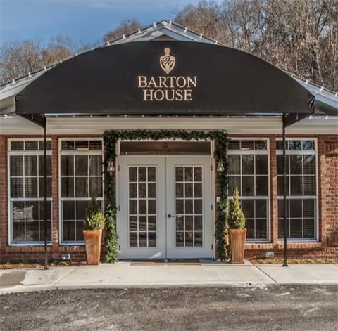 Front entrance of Barton House with a black awning displaying the name, double glass doors flanked by windows and potted plants.