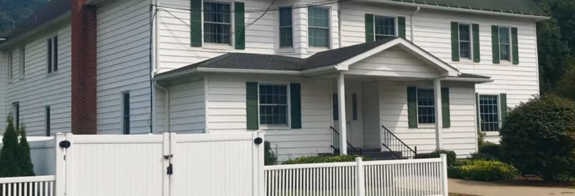 Front exterior of a two-story white house with green shutters, a small covered porch, and a white fence.