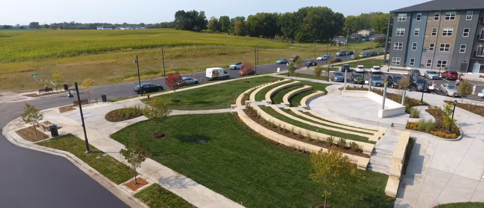 Aerial view of an outdoor terraced amphitheater and landscaped lawn next to a multi-story senior living building and parking lot with open fields beyond.