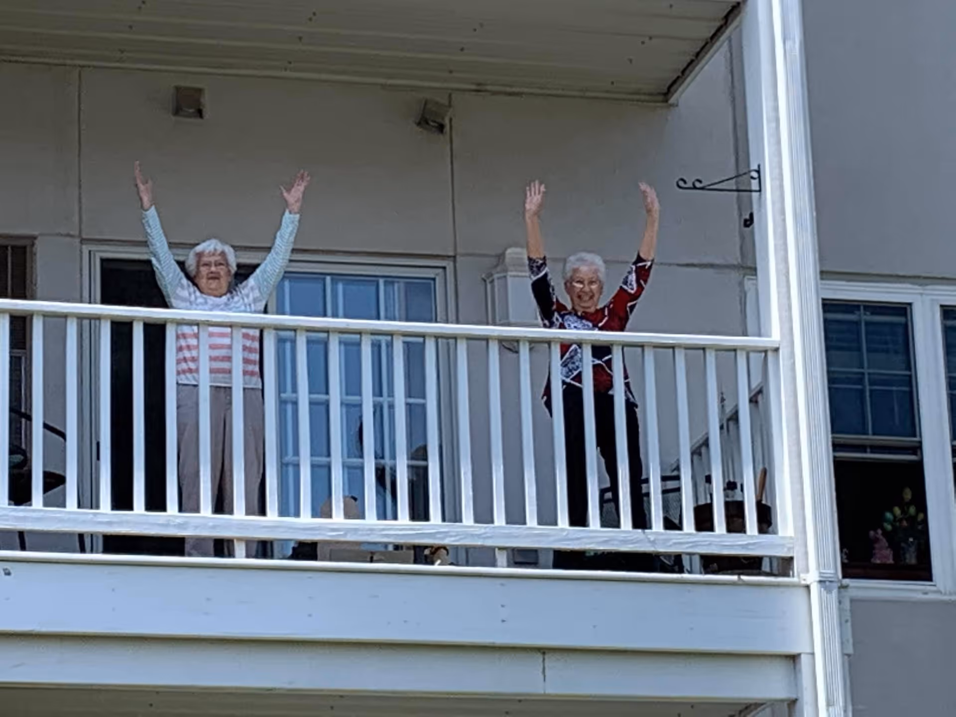 Two elderly women standing on a white balcony with their arms raised, smiling. The balcony has white railings and is attached to a building with windows and a door behind them.