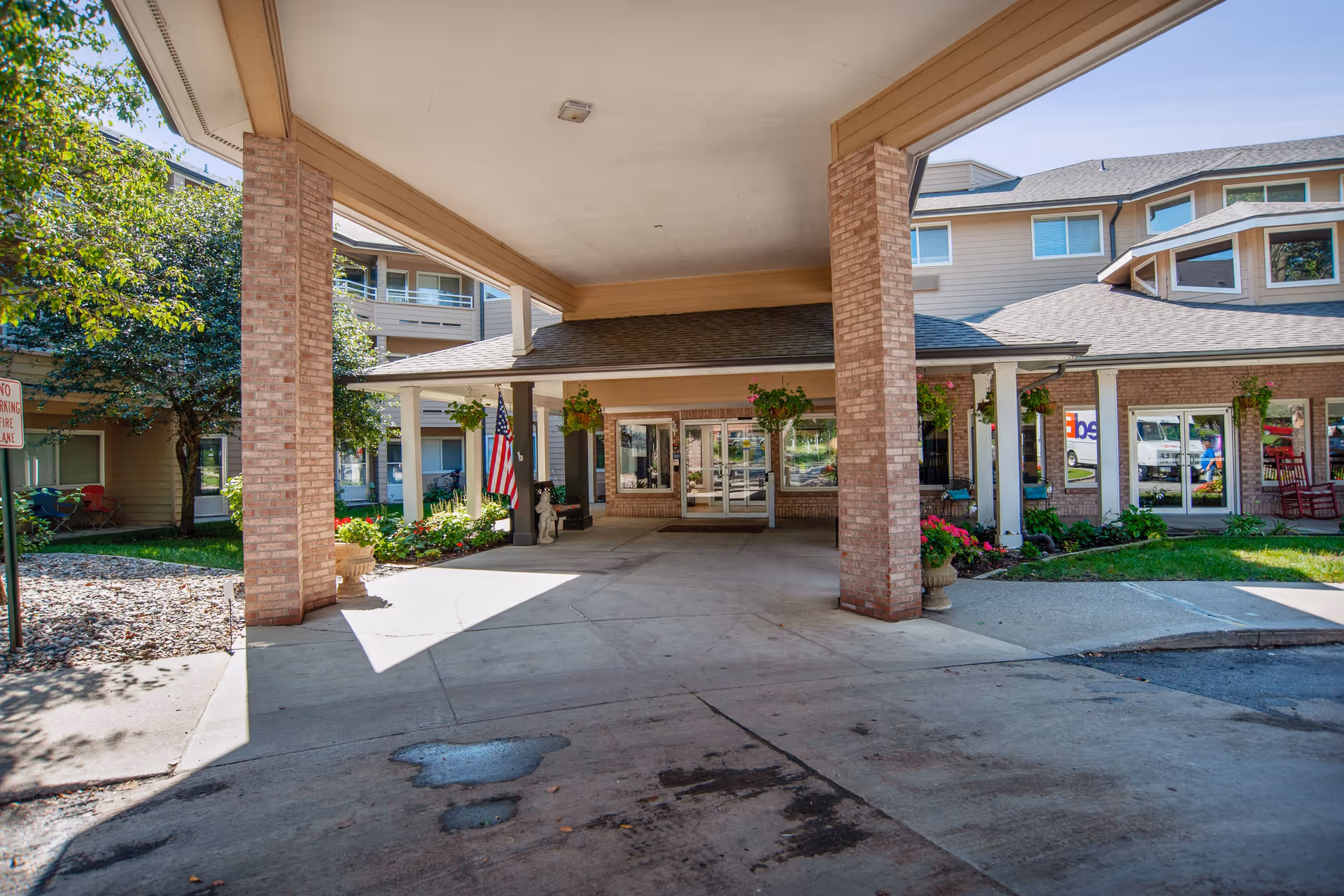 Covered entrance area of a senior living facility with brick pillars, an American flag, potted plants, and glass double doors leading inside. The building has beige siding and multiple windows, with greenery and flowers around the entrance.