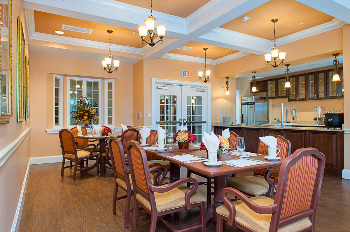 A well-lit dining room with wooden tables and cushioned chairs arranged neatly. The tables are set with white napkins, cups, glasses, and floral centerpieces. The room features warm beige walls, wooden flooring, and multiple hanging light fixtures. In the background, there is a kitchen area with dark wood cabinets, a refrigerator, and a countertop with a sink and appliances. Double glass doors are visible at the far end of the room.