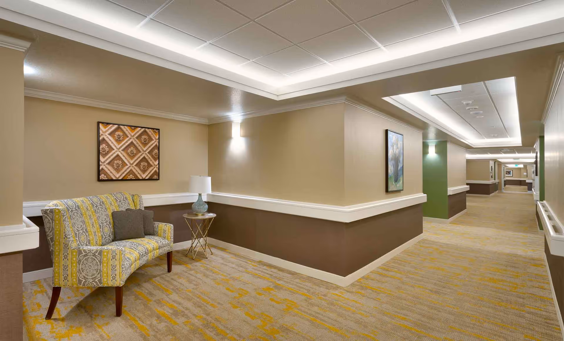 A well-lit hallway in a senior living facility with beige and brown walls, yellow patterned carpet, and ceiling lights. There is a patterned yellow and gray loveseat with two gray cushions and a small round side table with a lamp. Artwork is hung on the walls along the hallway.