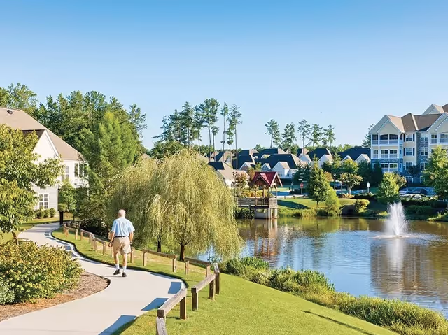 A person walking along a paved lakeside path with a fountain, gazebo and residential buildings in the background.