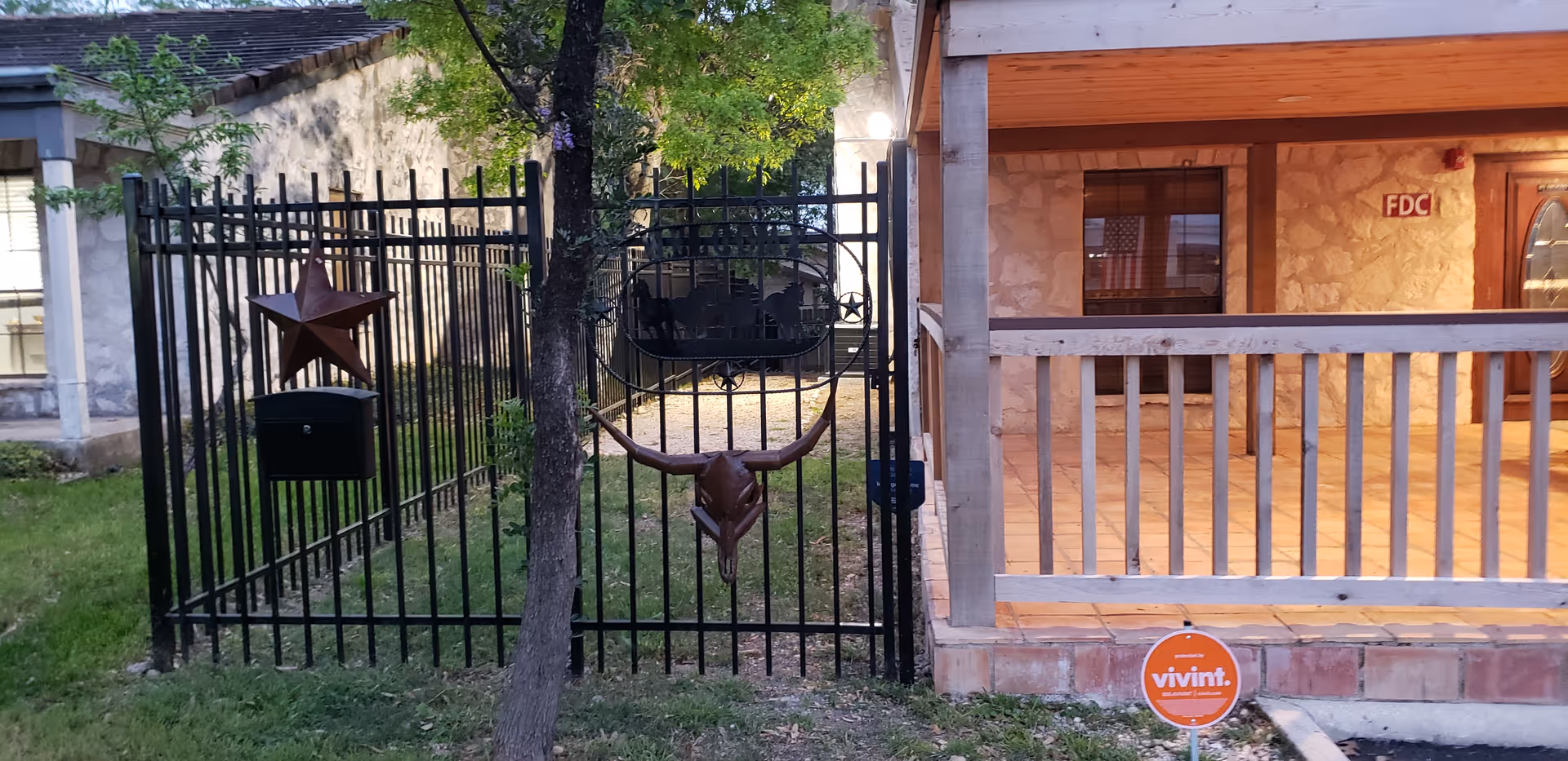 A gated entrance with a black metal fence featuring decorative elements including a star and a longhorn skull. Behind the gate is a stone building with a covered porch and wooden railing. A tree is in front of the gate, and a Vivint security sign is placed on the ground near the porch.