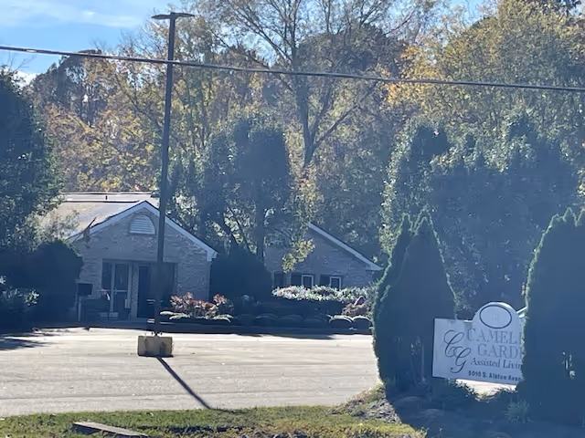 View of the front exterior of Camellia Gardens Assisted Living facility with a parking lot in the foreground, trees and shrubs surrounding the building, and a sign displaying the facility's name near the entrance.