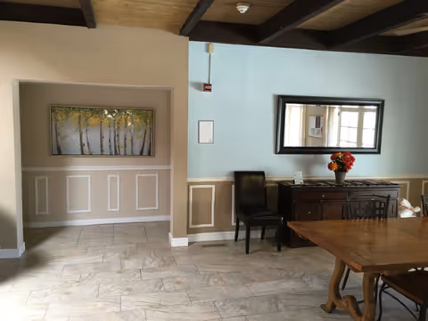 Interior view of a senior living facility showing a dining area with a wooden table and chairs, a dark wooden sideboard with a vase of flowers on top, a large mirror on the wall, and a painting of trees in an alcove. The walls are painted in beige and light blue with white trim, and the floor is tiled.