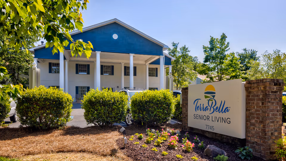 Exterior view of TerraBella Senior Living facility with a blue and white building featuring large white columns. There are green bushes and a flower bed in front of the building, along with a sign that reads 'TerraBella Senior Living 7745'. The sky is clear and blue.
