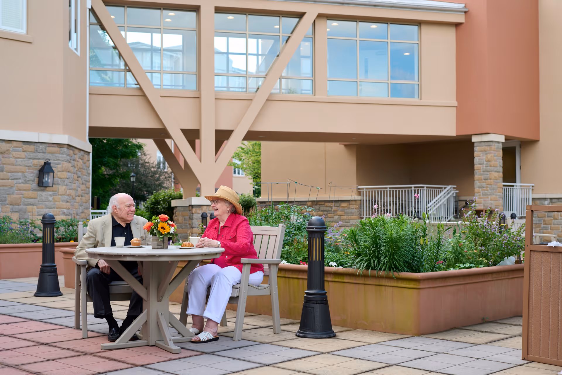 An elderly man and woman sitting at a round outdoor table in a courtyard area, enjoying a meal and conversation. The woman is wearing a hat and a bright pink jacket, while the man is dressed in a beige jacket. The courtyard features potted plants, stone walls, and a building with large windows in the background.
