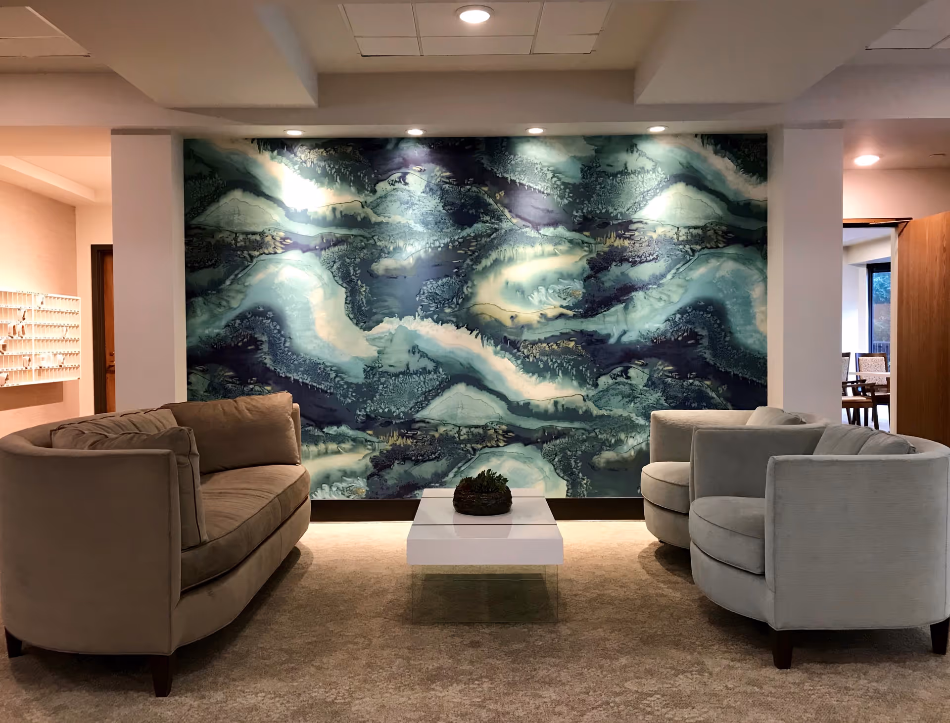 A cozy seating area with a beige sofa on the left and two light gray armchairs on the right, arranged around a low white coffee table with a small plant centerpiece. Behind the seating area is a large abstract wall art with swirling shades of blue, green, and white, illuminated by spotlights from above. The space has a warm, inviting atmosphere with carpeted flooring and soft lighting.