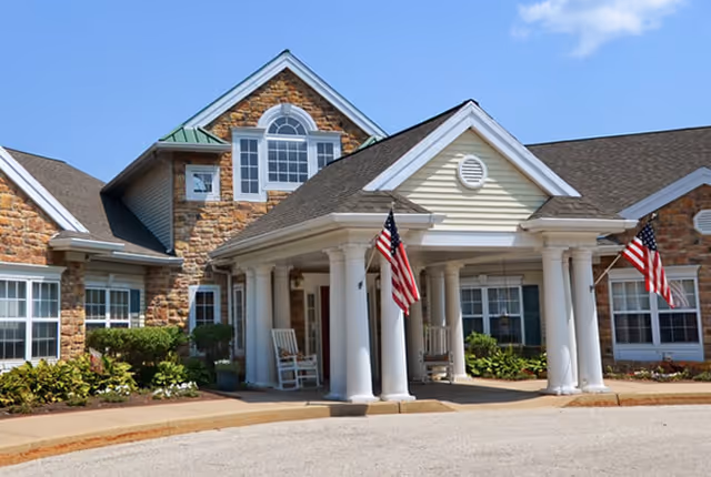 Front exterior view of a senior living facility named Park Creek Place featuring a stone and siding facade, a covered entrance with white columns, two American flags, rocking chairs on the porch, and a clear blue sky.