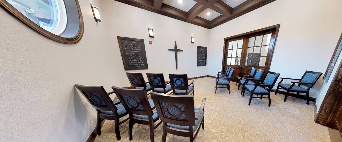 A small chapel or meditation room with wooden chairs arranged in rows facing a wall with a wooden cross and two framed plaques. The room has beige carpet, white walls, wooden trim, and a coffered ceiling with recessed lighting. There is a round window on the left wall and double wooden doors with glass panes on the far side.