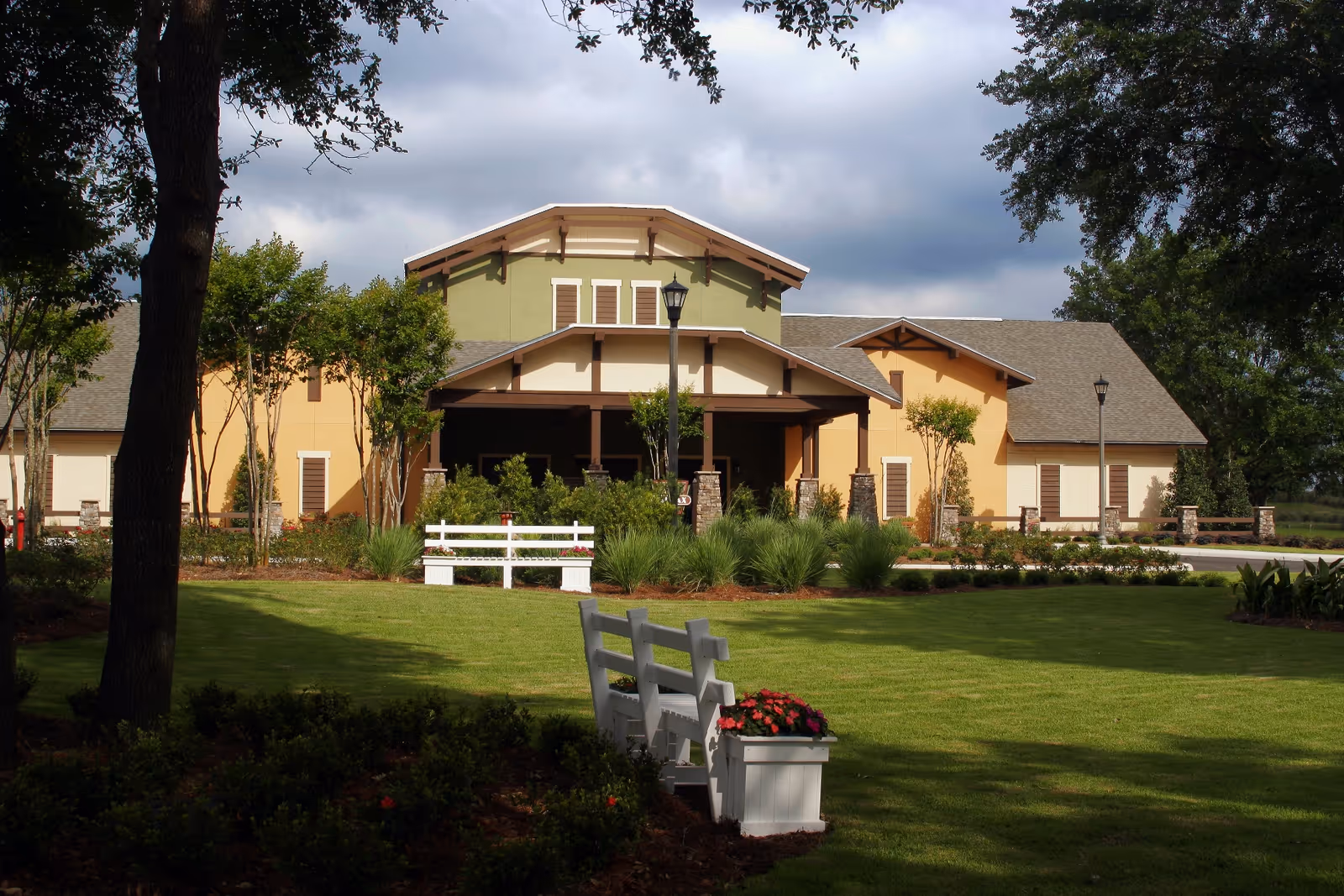 A large, single-story building with a green and beige exterior surrounded by well-maintained landscaping including trees, bushes, and a green lawn. There are white benches and flower planters on the lawn in front of the building. The sky is partly cloudy.