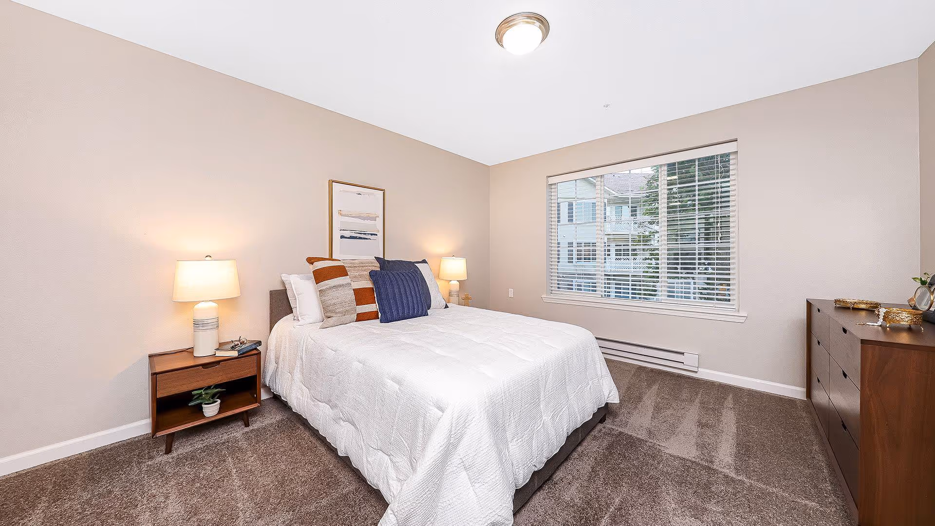 A neatly arranged bedroom with a bed covered in a white quilt and multiple pillows in various colors. There are two bedside tables with lamps on each side of the bed. A framed artwork hangs above the bed. The room has beige walls, a window with blinds, and a brown carpeted floor. A wooden dresser is positioned against the right wall.