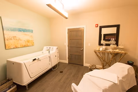 A serene therapy or spa room featuring a white hydrotherapy tub on the left, a massage table covered with a white sheet on the right, a wooden door in the center background, a decorative table with seashells and a mirror above it on the right wall, and a calming beach-themed painting on the left wall.