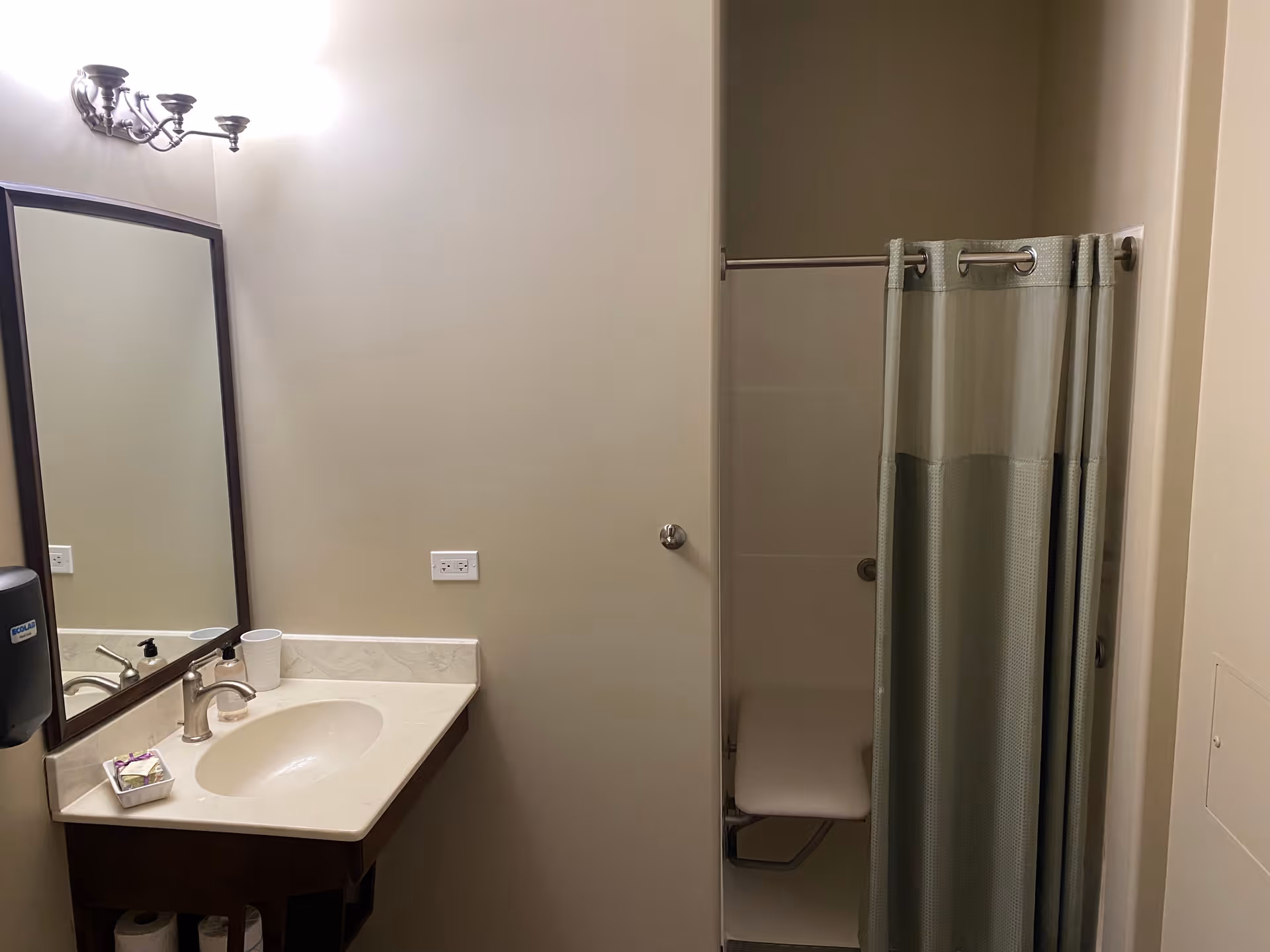 A clean bathroom with a sink and countertop on the left side, a large mirror above the sink, a wall-mounted soap dispenser, and a small tray with toiletries. On the right side, there is a shower area with a curtain and a foldable shower seat inside.