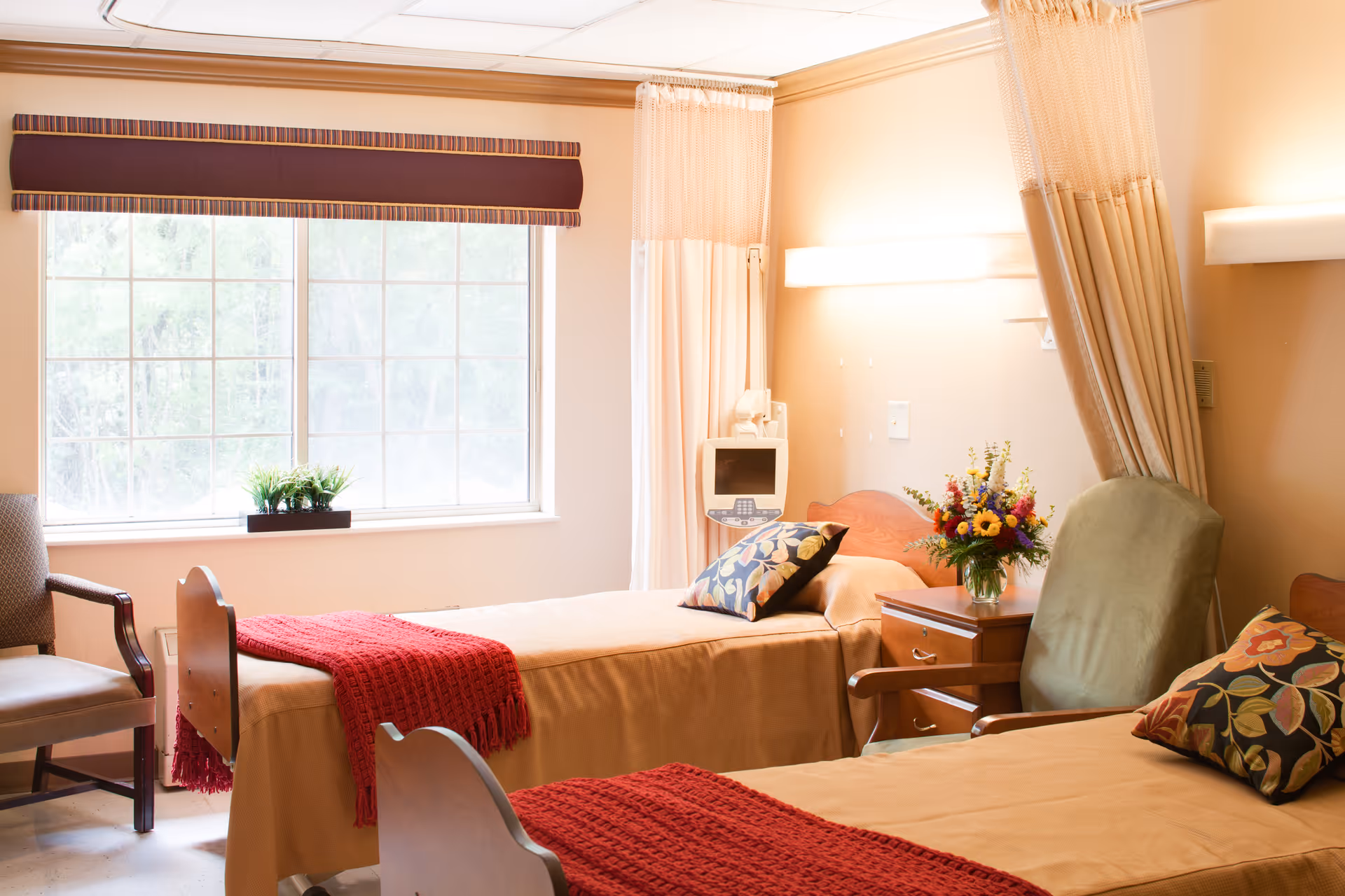 Bright shared senior care room with two beds, chairs, a window, and a bedside table with flowers.
