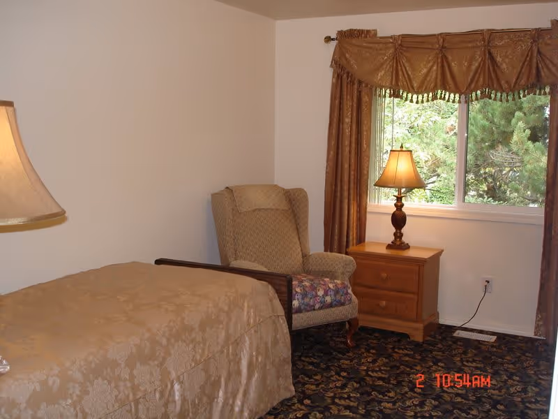 A cozy bedroom with a single bed covered in a beige patterned bedspread, a cushioned armchair with a floral seat cushion, a wooden nightstand with a lamp, and a window with brown curtains showing greenery outside.