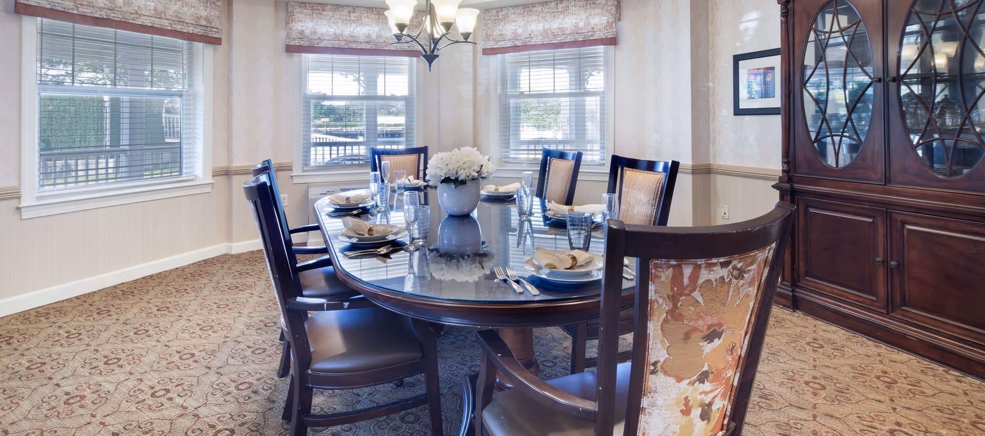 A well-lit dining room with a large oval wooden table set for six people. The table has plates, glasses, napkins with rings, and a white flower centerpiece. There are six wooden chairs around the table, one with a floral patterned backrest. The room has three windows with blinds and valances, a chandelier overhead, patterned carpet, and a dark wooden china cabinet with glass doors displaying glassware.