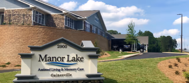 A stone sign reading "Manor Lake Assisted Living & Memory Care - Gainesville" sits on a lawn in front of the facility building and driveway under a blue sky.