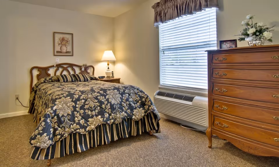 A cozy bedroom in a senior living facility featuring a bed with a floral and striped bedspread, a wooden headboard, a nightstand with a lamp and phone, a window with blinds and a valance, and a wooden dresser with decorative items on top.