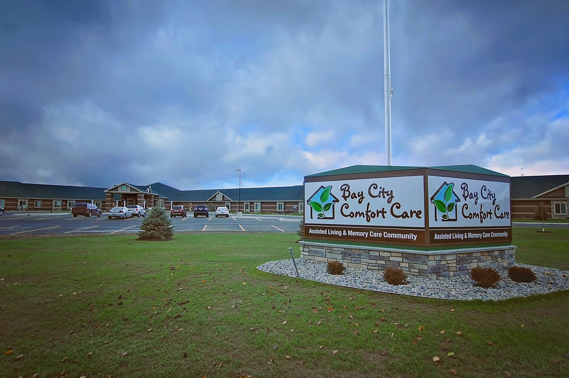 Exterior view of Bay City Comfort Care, an assisted living and memory care community, showing a large building with a parking lot and several cars parked. A prominent sign with the facility's name and logo is displayed on a stone base in the foreground, with a cloudy sky overhead.