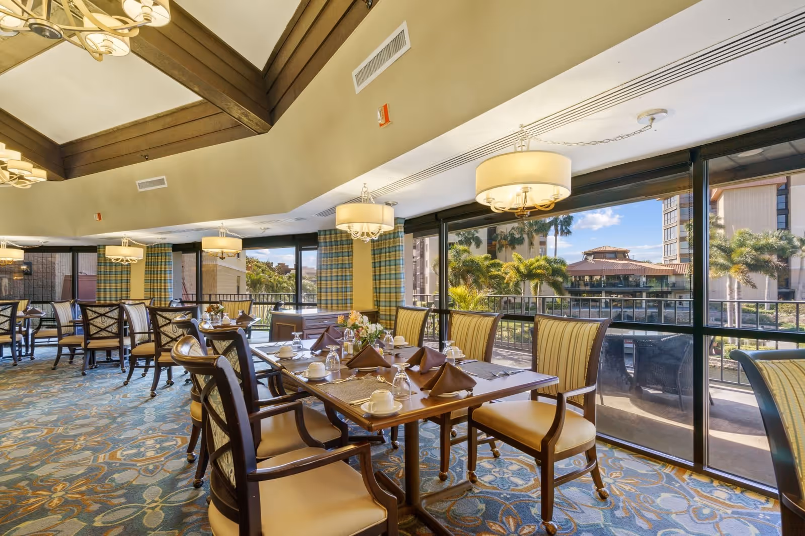 A bright and spacious dining room with large windows offering a view of palm trees and buildings outside. The room features a high ceiling with wooden beams, multiple round chandeliers, and tables set with brown napkins, white cups, glasses, and cutlery. The carpet has a blue and beige floral pattern, and the chairs have wooden frames with beige cushions and striped upholstery.