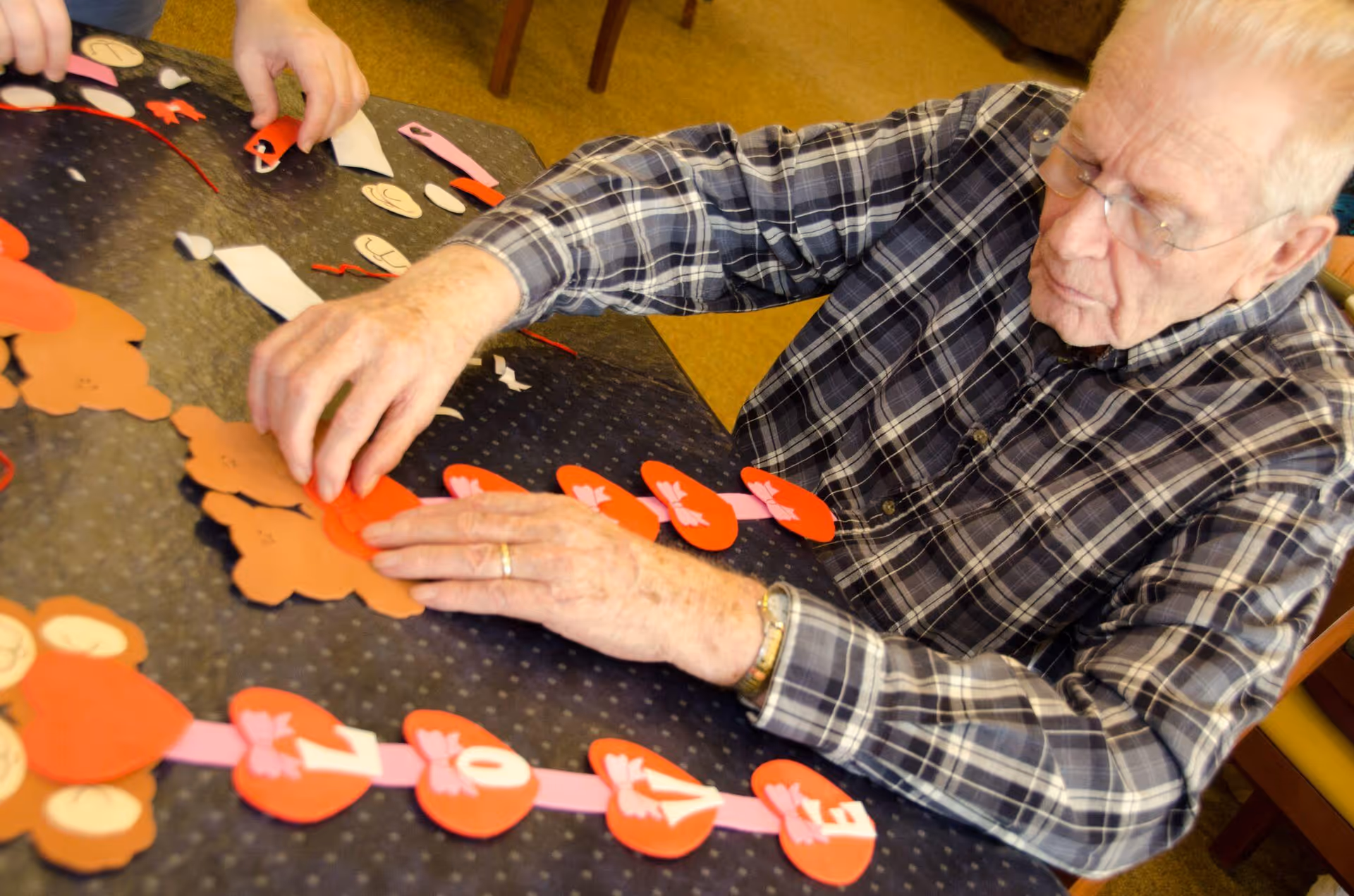 An elderly man wearing glasses and a plaid shirt is seated at a table working on a craft project involving red heart-shaped cutouts and brown bear shapes. Another person's hands are also visible assisting with the craft on the table.