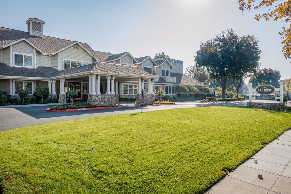 Exterior view of a senior living facility with a large green lawn in front, a covered entrance with stone pillars, and a sign that reads 'Sunrise Senior Living'. Trees and shrubs surround the building and the area appears well-maintained under a clear sky.