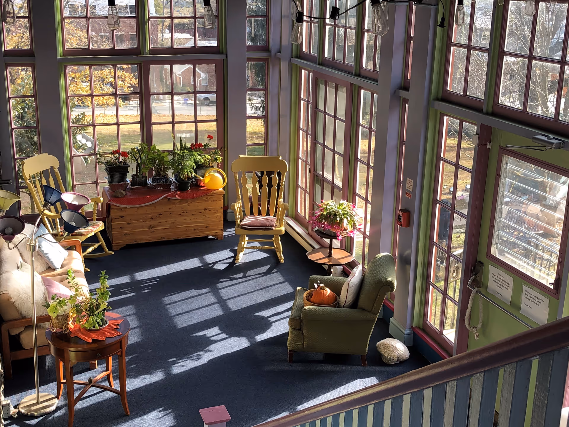 A bright sunroom with large windows letting in natural light, featuring a green armchair with a pumpkin on it, two yellow wooden rocking chairs, a wooden chest with potted plants on top, a small round table with a plant, a beige sofa with cushions, and a floor lamp with multiple adjustable heads. The room has a blue carpet and light green and purple painted walls and window frames.