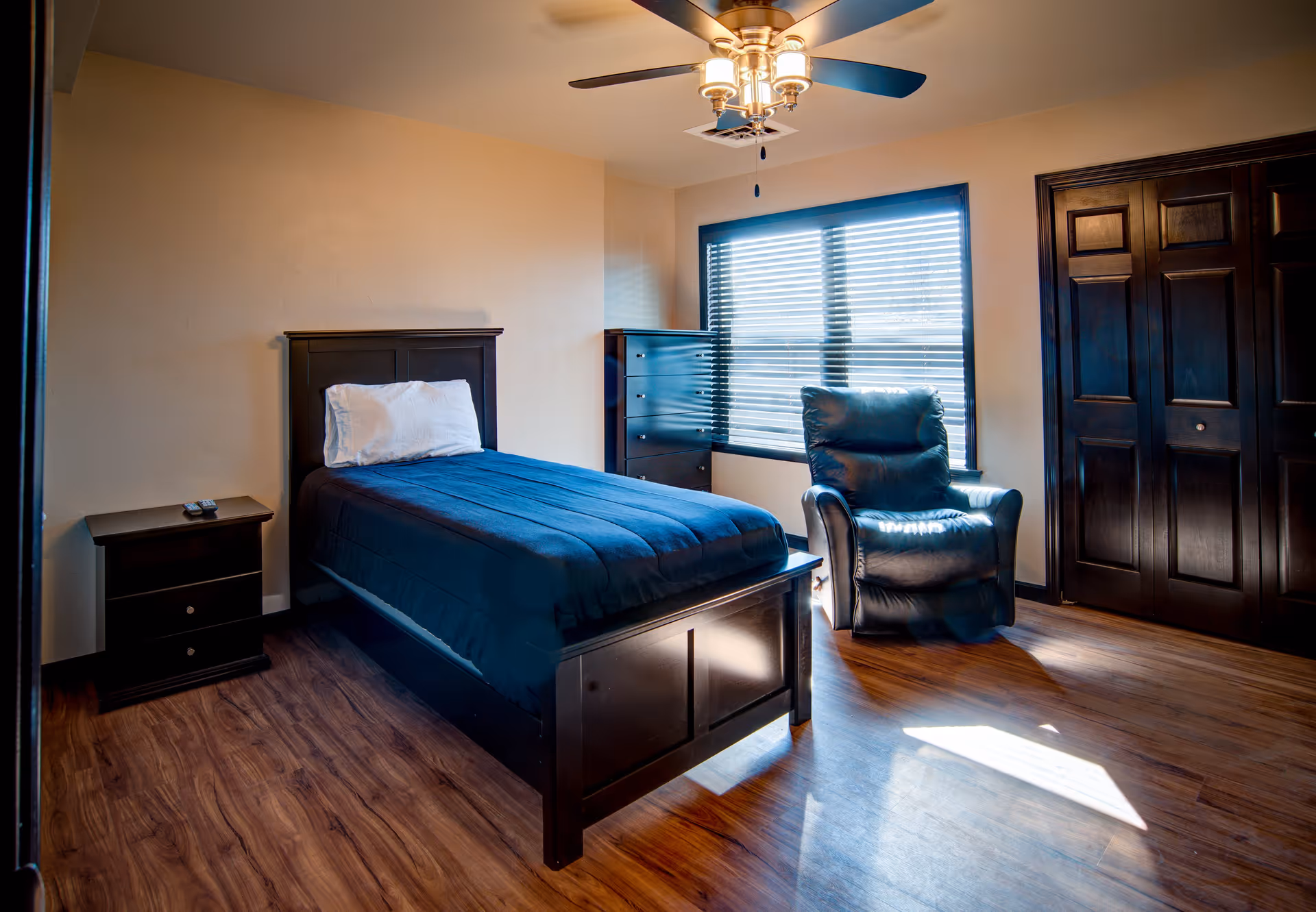 A bright bedroom with a single bed covered with a dark blue comforter and a white pillow. Next to the bed is a small dark wooden nightstand with two drawers and a remote control on top. Across from the bed is a black leather recliner chair positioned near a window with closed blinds. A tall dark wooden dresser is placed against the wall near the window. The room has wooden flooring, beige walls, a ceiling fan with lights, and dark wooden closet doors.